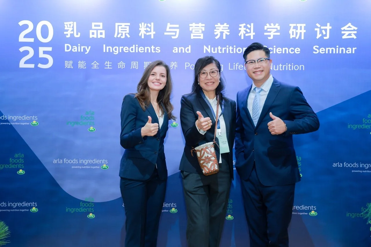 Three people in business attire standing in front of a blue conference backdrop, smiling and giving thumbs up. The backdrop has Chinese and English text about a dairy ingredients and nutrition science seminar.