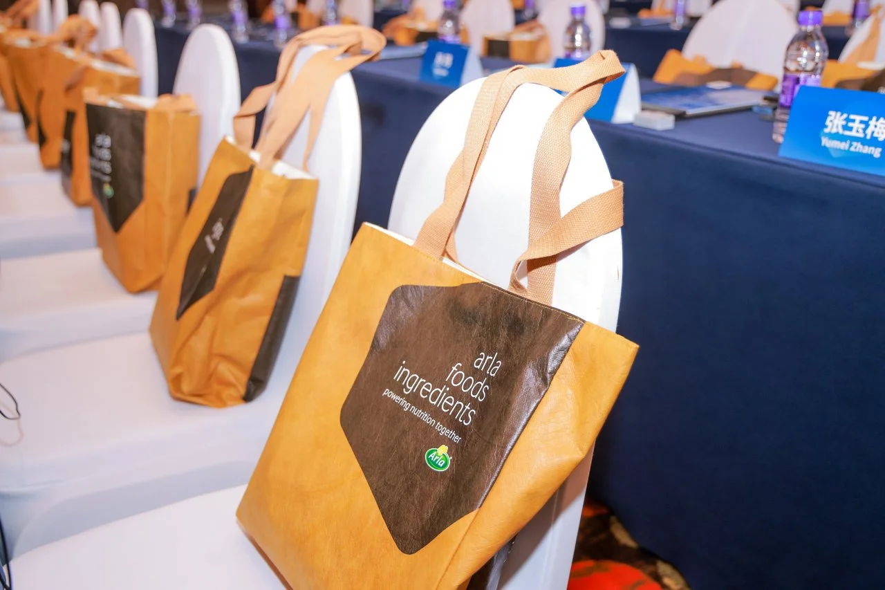 A row of white chairs with tan tote bags hanging on them, each bag labeled "arla foods ingredients" and sitting in front of a meeting table with water bottles and name tags.