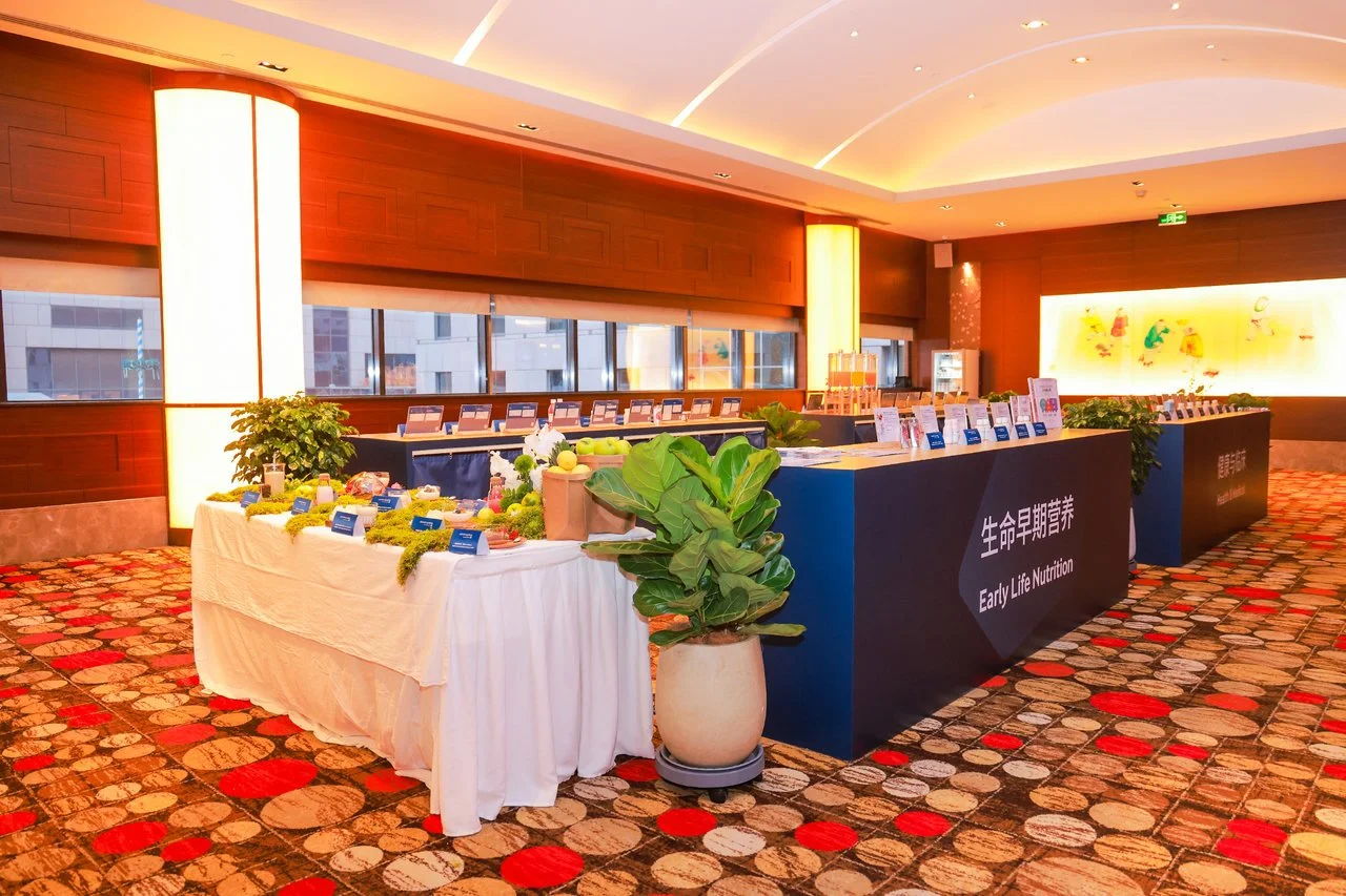 Indoor display of healthy foods and nutritional products, featuring a table with fresh vegetables, fruits, and labels, with a backdrop of a modern, well-lit room.