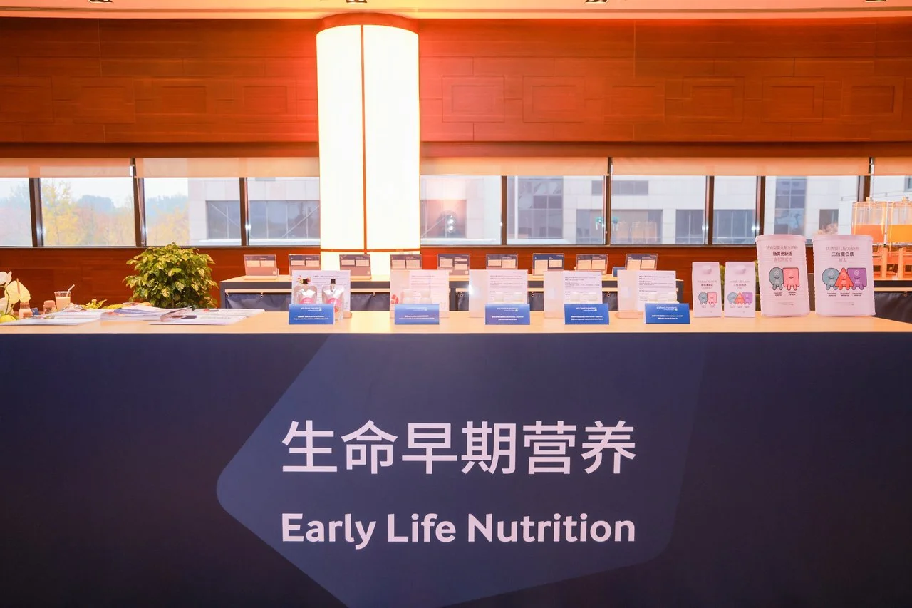 Table with informational brochures and pamphlets about early life nutrition, set against a wooden-paneled wall with large windows, in a well-lit conference or exhibition area.