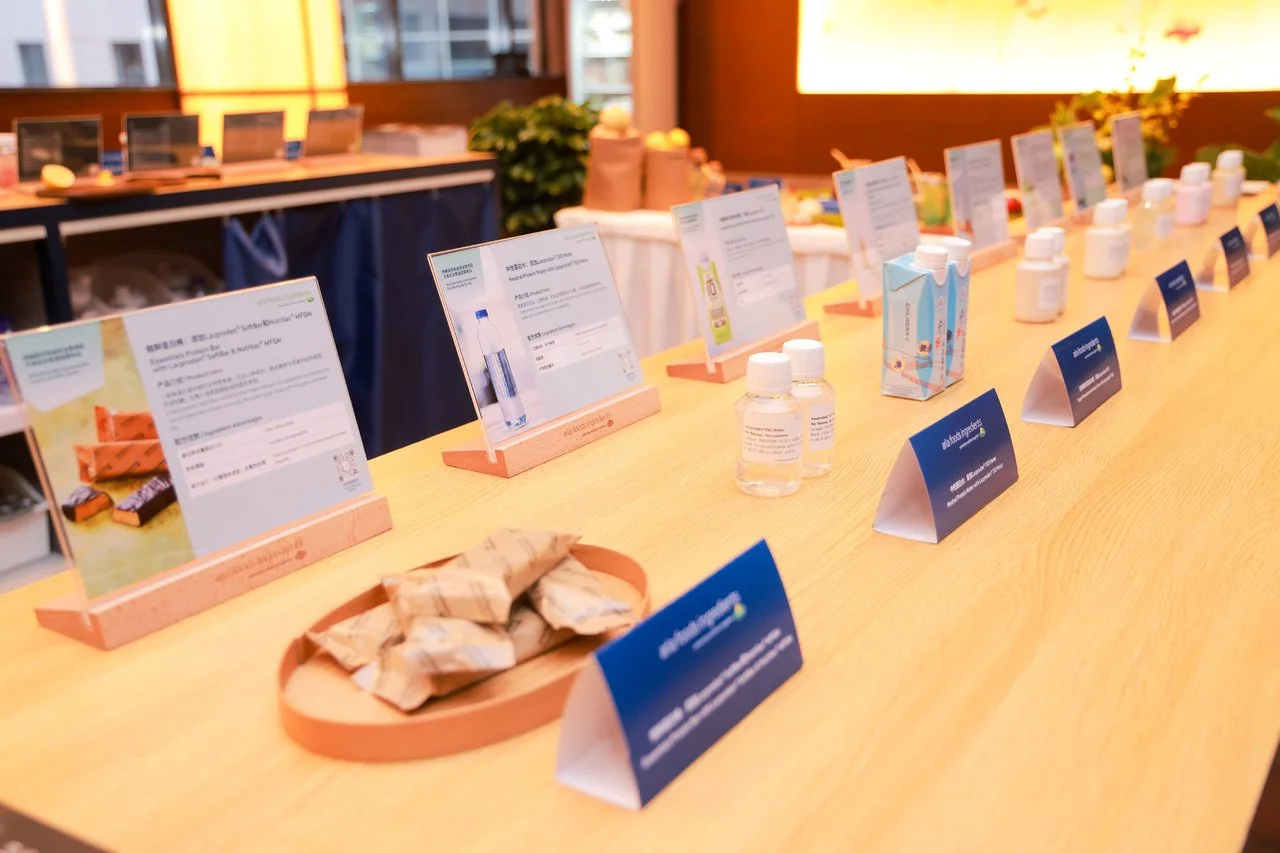 Display table with various informational cards and product samples, including small bottles and packaged items, in a well-lit room with wooden accents and decorative plants.