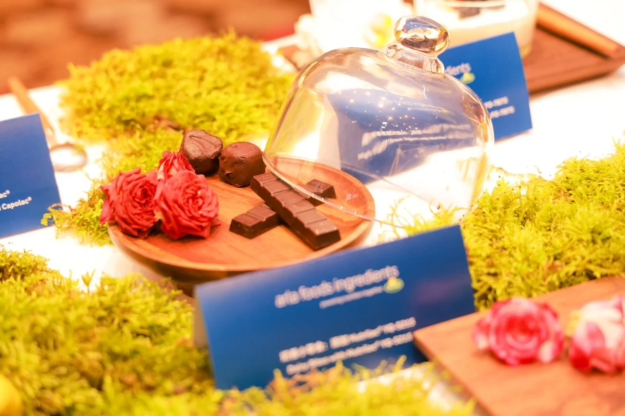 Chocolate truffles, chocolates, red roses, and decorative moss on a display table with blue informational cards, covered with a glass dome.