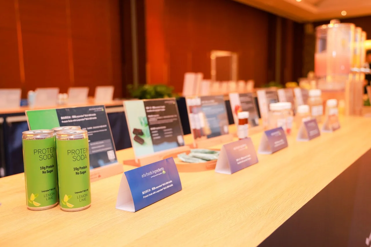 Display table with multiple product samples and informational cards at a promotional event, featuring cans of protein soda labeled lemon lime in the foreground.