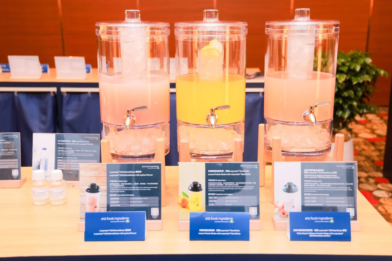 Three large clear dispensers filled with pink, yellow, and pink liquids, possibly beverages, on a table at an event. Small bottles and informational cards are placed in front of the dispensers.