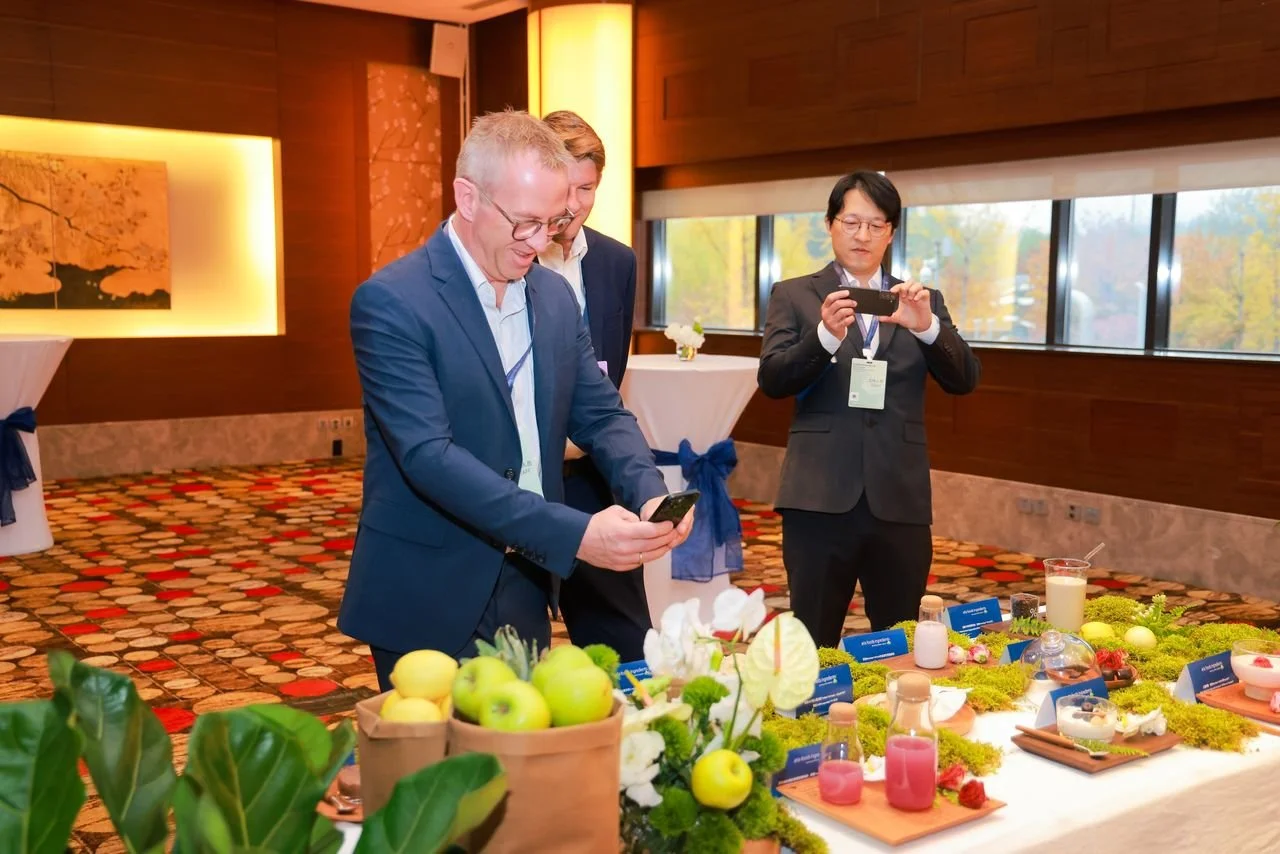 Three men in business suits taking photos of a table with various food items and decorative plants inside a well-lit room with large windows and fall outdoor scenery.