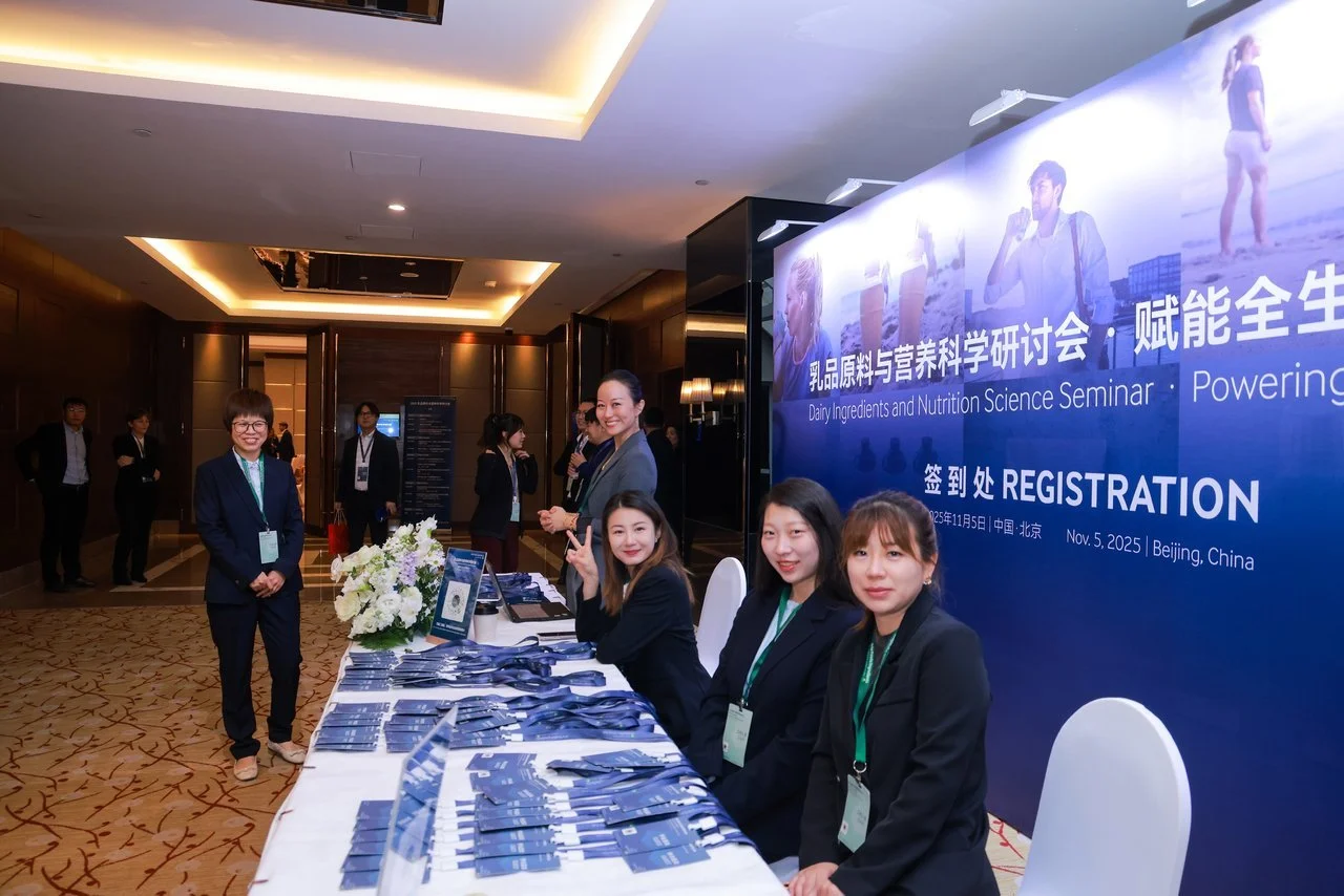 Registration desk at the Dairy Ingredients and Nutrition Science Seminar in Beijing, China on November 5, 2025. Several women are sitting and standing behind the desk, smiling and wearing formal attire, with a large blue event banner behind them.