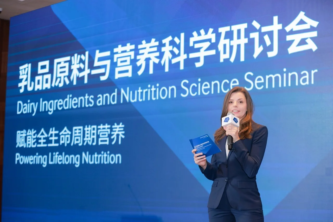 A woman in a navy blazer standing in front of a large blue screen with white text. She is speaking into a microphone and holding a small booklet. The screen displays text in Chinese and English about a seminar on Dairy Ingredients and Nutrition Science, with a subtitle mentioning powering lifelong nutrition.