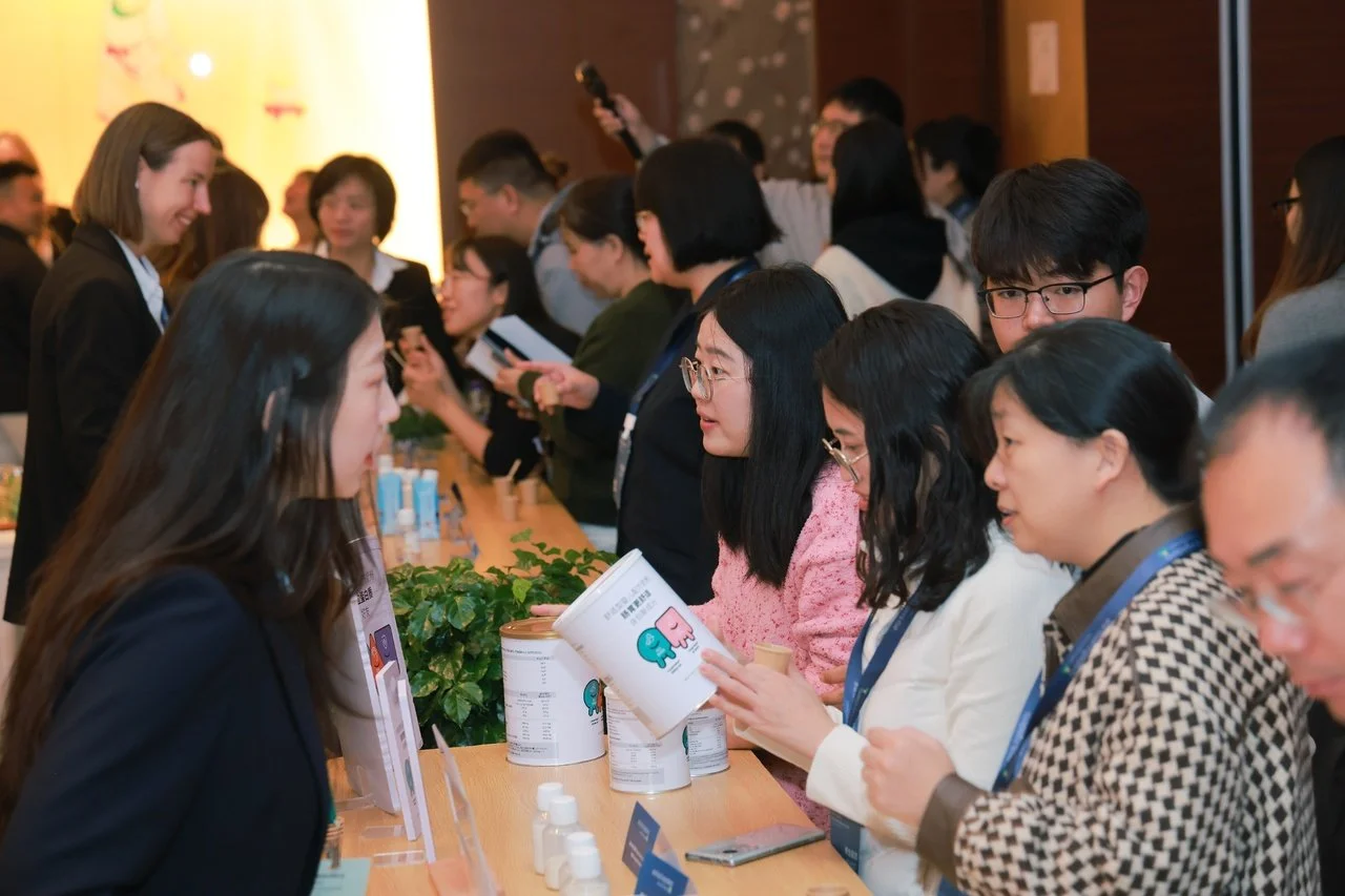 People at an event or conference, standing at a long table with name tags and informational materials, engaging in conversations.
