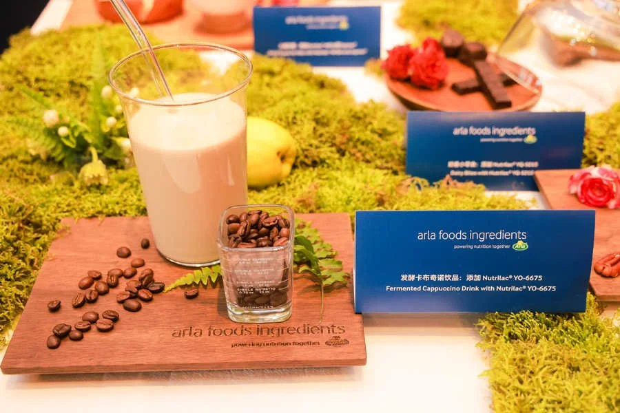 A glass of fermented cappuccino drink with a straw next to a small glass filled with coffee beans on a wooden board, with blue Arla Foods ingredients signage in the background.
