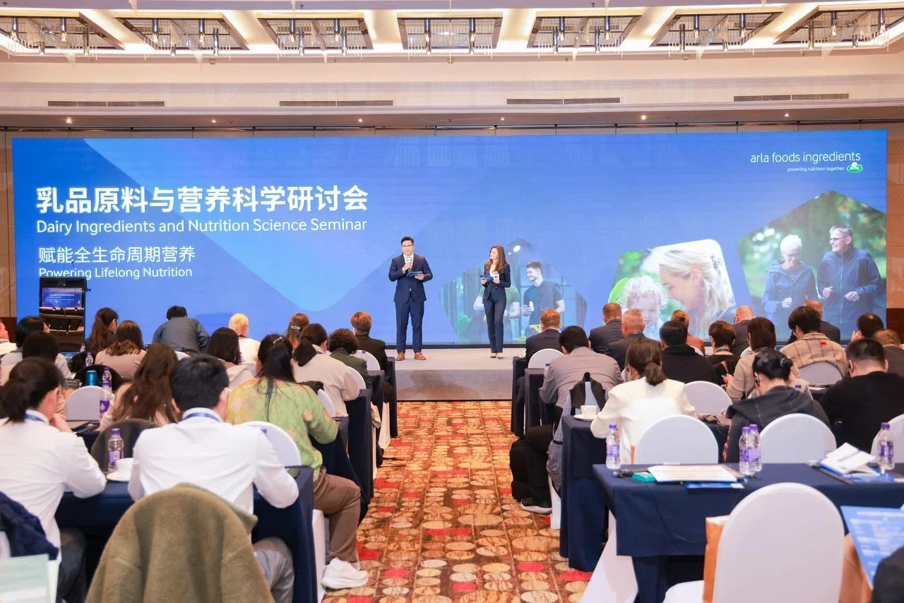 A large conference hall with people attending a seminar titled 'Dairy Ingredients and Nutrition Science Seminar' on a blue screen. Two presenters, a man and a woman, are speaking on stage. The audience is seated at tables, some taking notes, with bottles of water on the tables.
