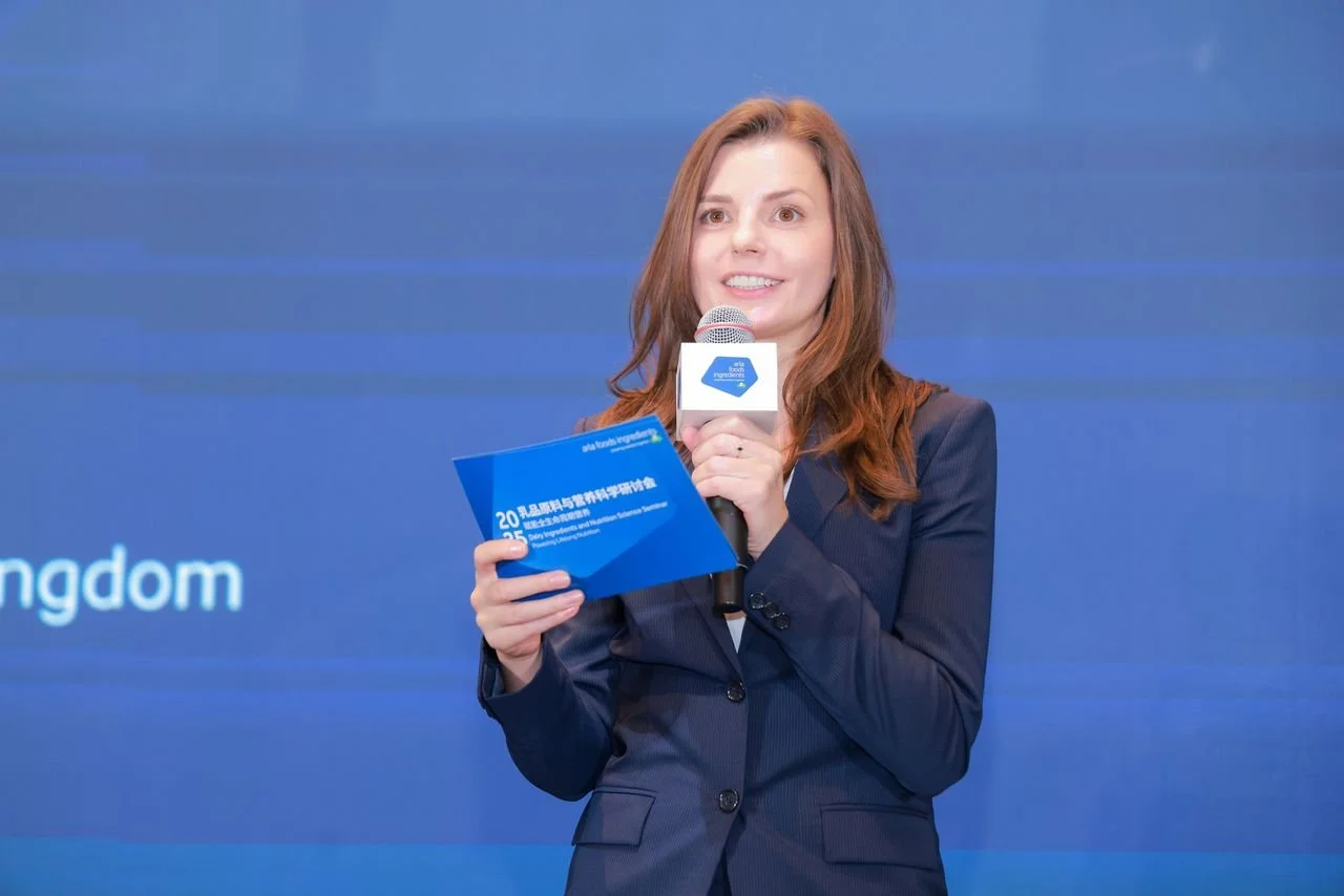 Woman with brown hair in a blazer holding a microphone and a blue booklet, standing against a blue background.