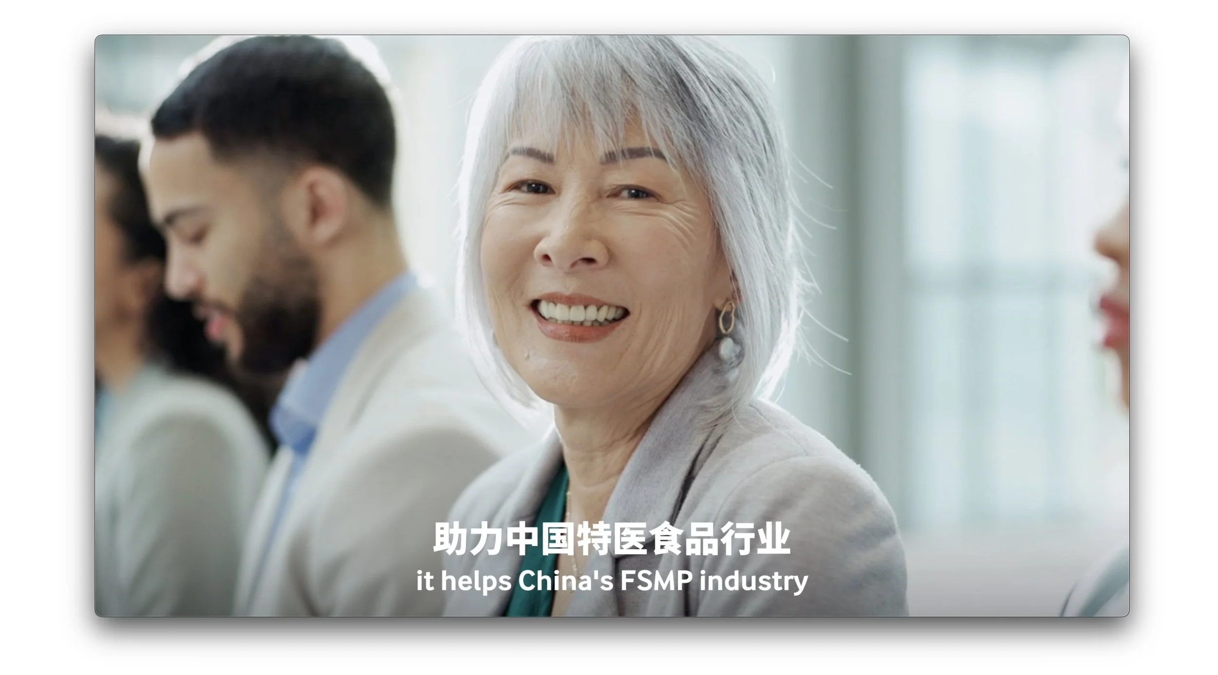 Smiling elderly Asian woman with gray hair, wearing earrings, at a professional conference or meeting, with other people in the background, and Chinese and English text mentioning support for China's FSMP industry.