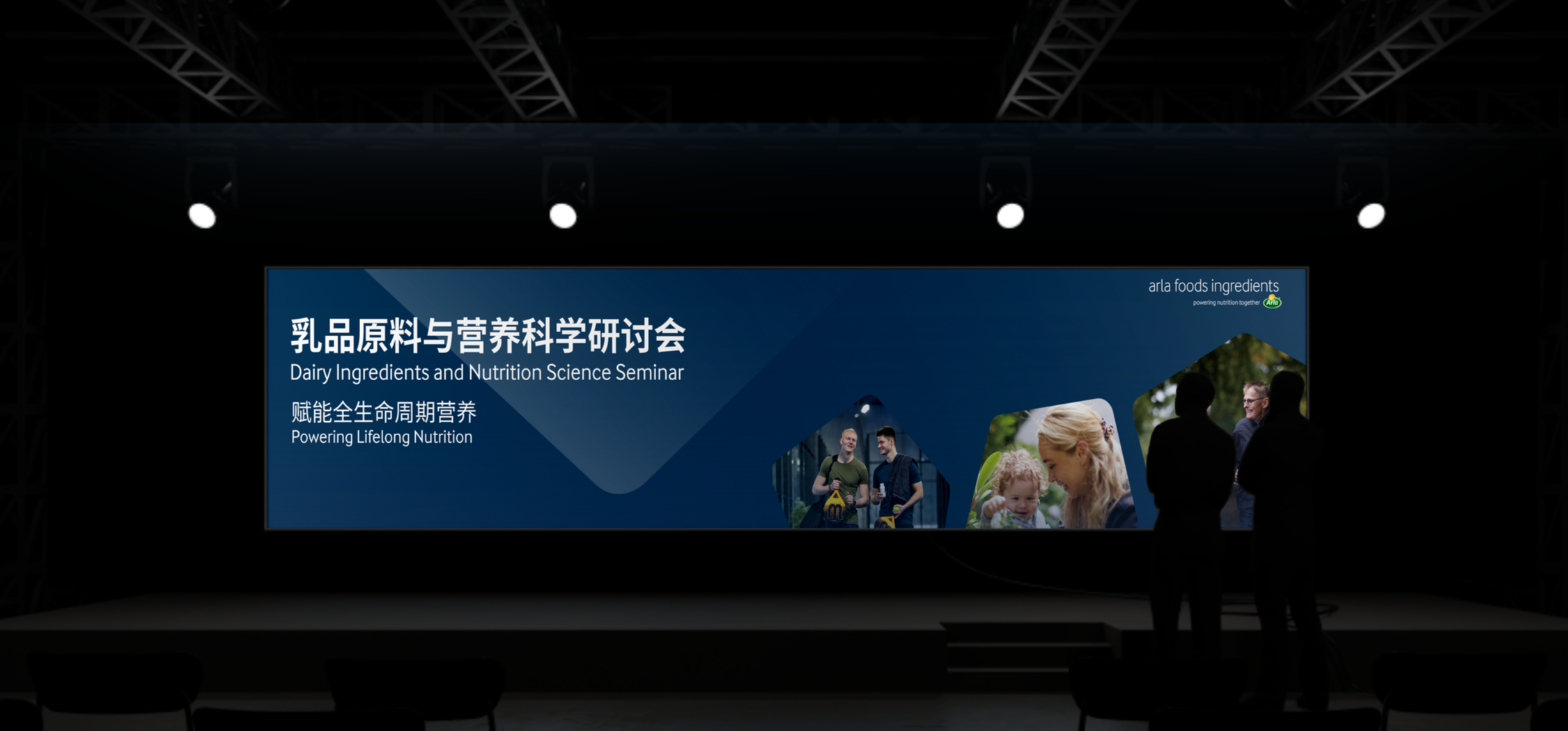 Large conference screen displays text and images about a Dairy Ingredients and Nutrition Science Seminar. The screen shows a blue background with white Chinese and English text, and photos of people talking and smiling. Three silhouetted people stand in front of the screen. Stage lights are visible overhead.