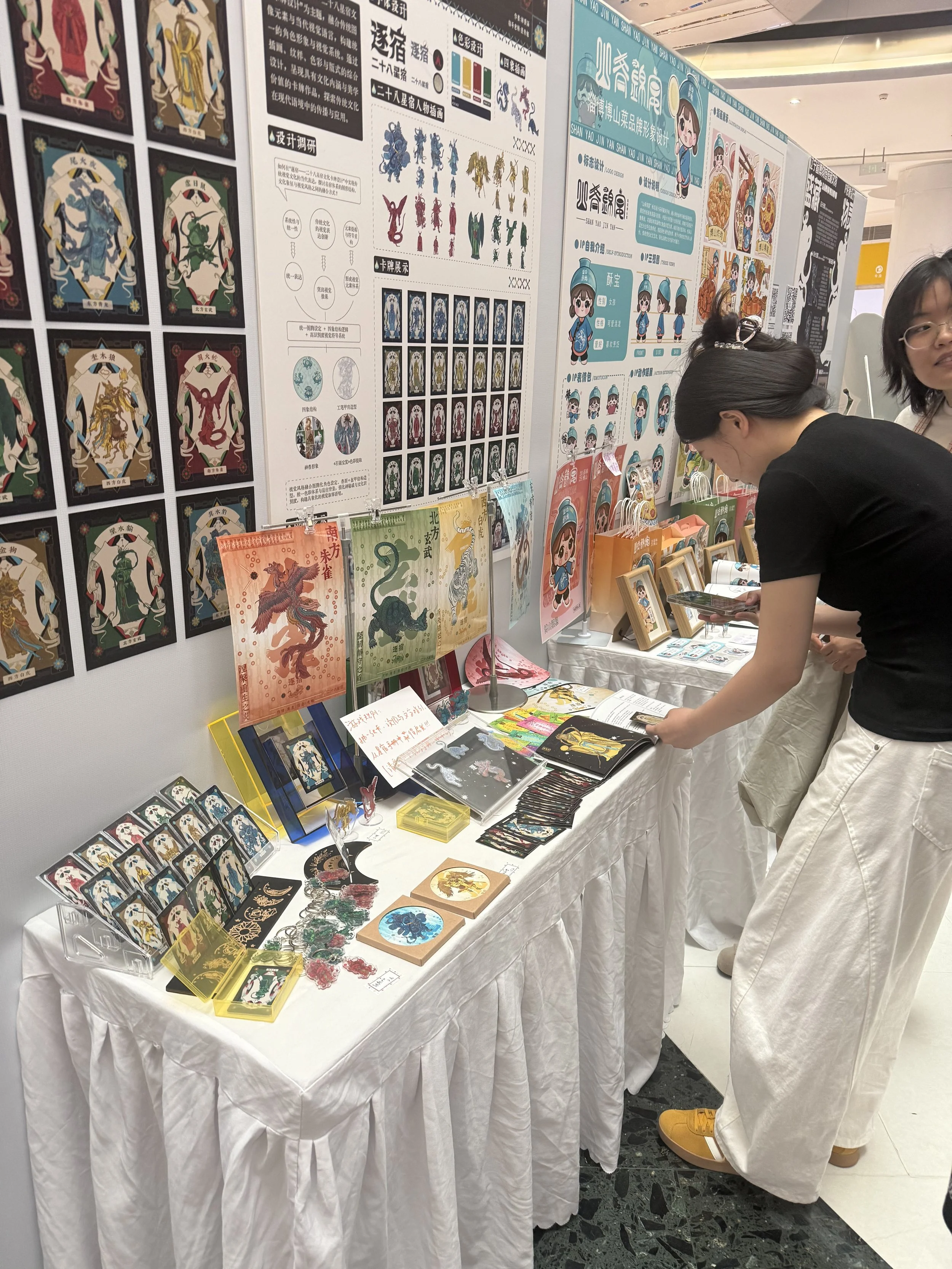 A display booth with various colorful prints, cards, and merchandise related to Chinese mythology or folklore, with text in Chinese characters. Two women are browsing the items. The background features posters with illustrations of characters and creatures.