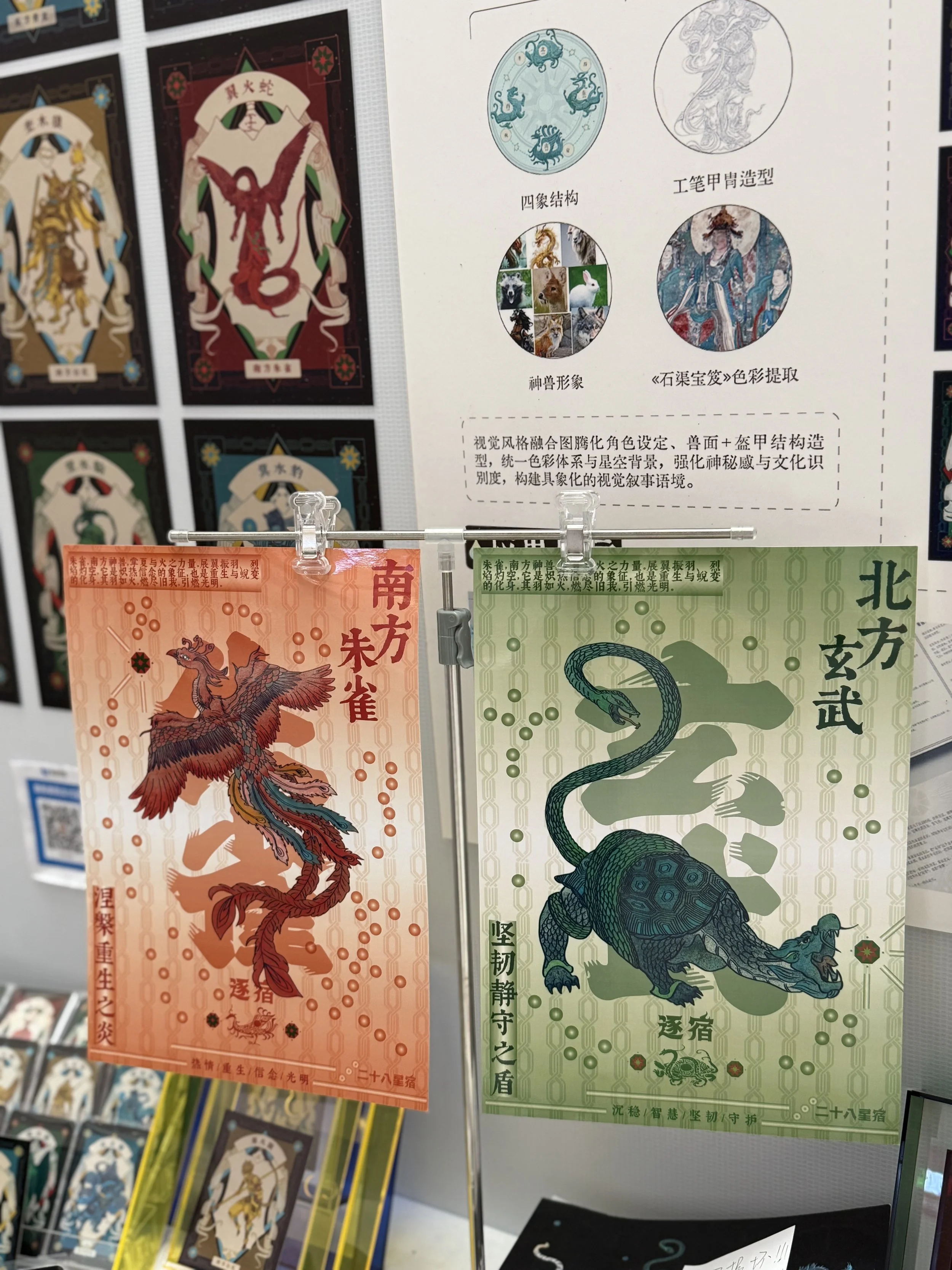 Two colorful tarot cards hanging on a display, one orange with a phoenix illustration and Chinese characters, and one green with a tortoise and snake illustration with Chinese characters, surrounded by informational posters and other cards.