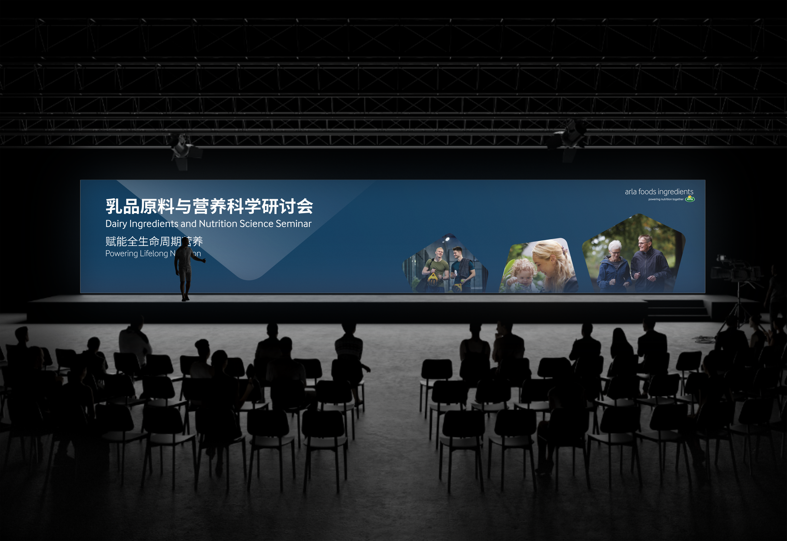 A presenter stands on a stage in front of a large digital screen displaying information about a Dairy Ingredients and Nutrition Science Seminar, with a row of chairs and seated conference attendees in a dark auditorium.