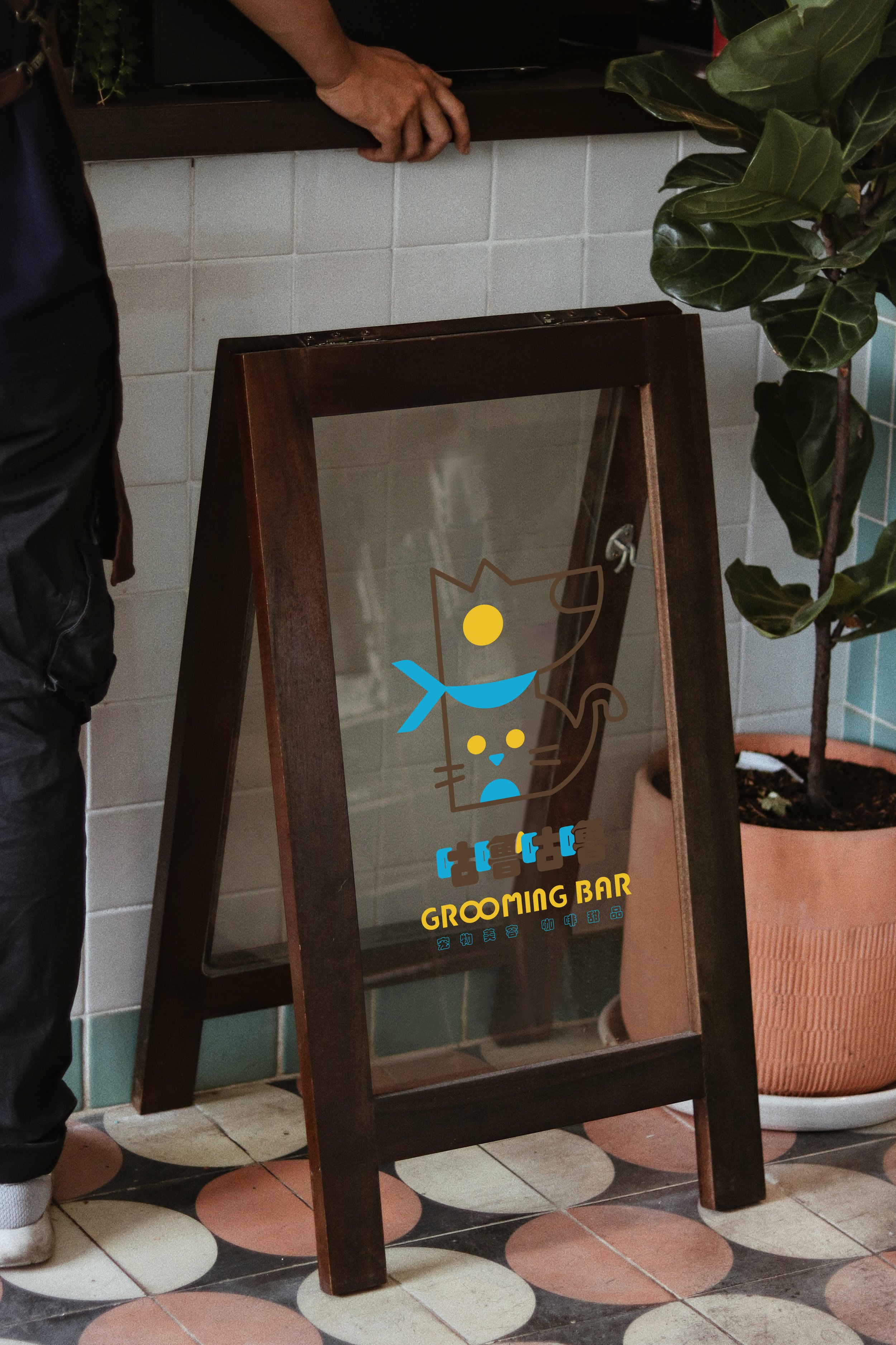 A wooden A-frame signboard with a glass front displays a logo of a stylized cat with a blue scarf and the words 'Grooming Bar' in yellow text. The signboard is positioned on a tiled floor next to a potted plant with large green leaves.