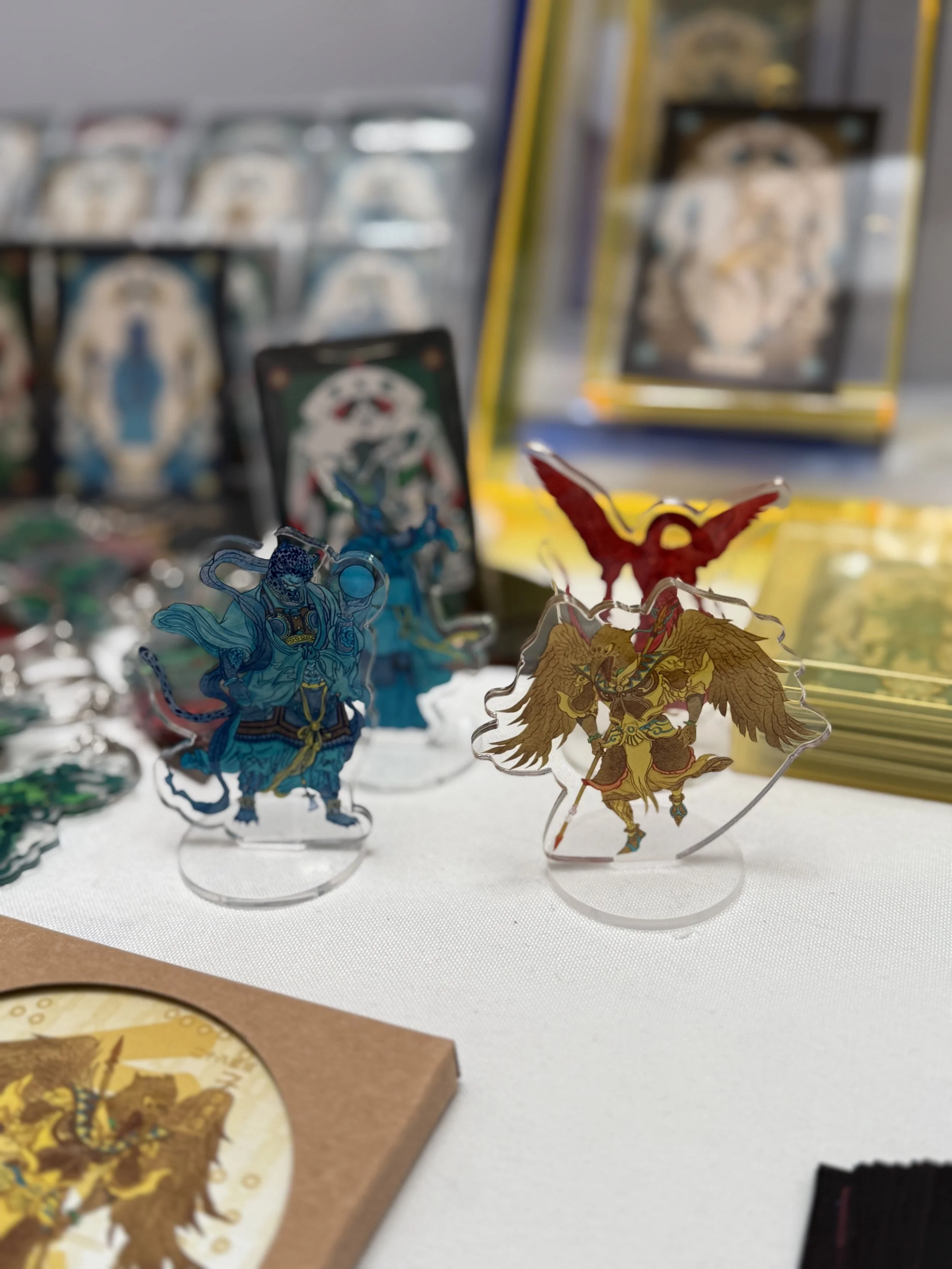 Colorful anime-style character standees on a table at a convention or store, with manga or art prints in the background.
